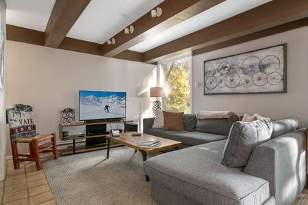 A cozy living room features a gray L-shaped sofa, a wooden coffee table, a flat-screen TV displaying a skiing scene, and a large window allowing natural light to fill the space.