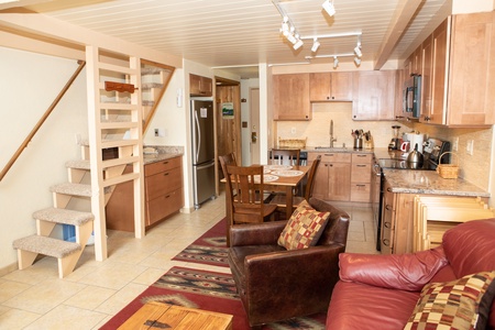 A small kitchen area with wooden cabinets, stainless steel appliances, a table with chairs, and a cozy living space with a red leather sofa and a wooden stairway leading to a loft.