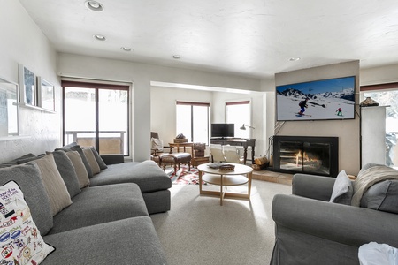 A modern living room features a gray sectional sofa, armchairs, a fireplace, and a wall-mounted TV displaying a skiing scene. Large windows provide ample natural light.