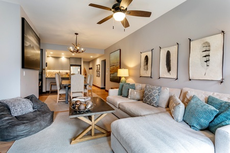 A modern living room with a beige sectional sofa, blue and gray pillows, a ceiling fan, and wall art. A dining area with a table and chairs is in view, leading to a kitchen with recessed lighting.