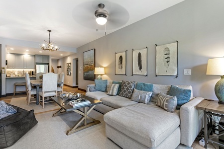 Modern living room with sectional sofa, decorative pillows, coffee table, and wall art. Dining area with table and chairs in the background, adjacent to a kitchen with bar seating.