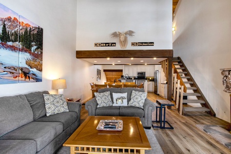 An interior shot of a modern living room with a gray sofa, wooden coffee table, stairs, and a kitchen in the background. Decor includes a cow skull and nature-themed photographs.