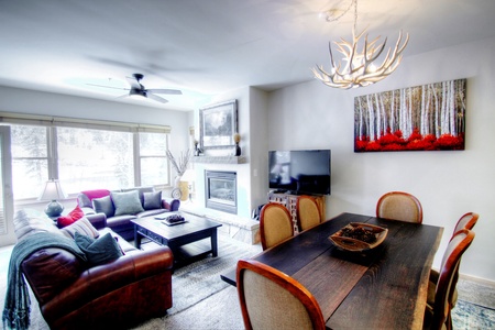 A modern living room with a leather sofa, a fireplace, a large window, a TV on a stand, and a dining table with six chairs. Antler chandelier and forest artwork enhance the decor.