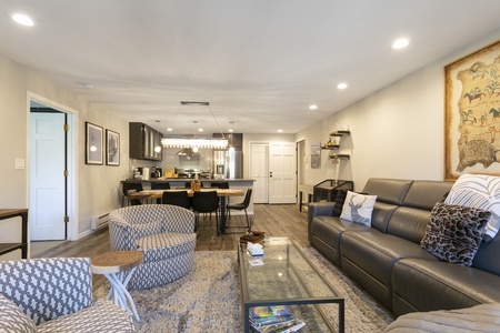 A modern living room with a gray sofa, patterned armchairs, and a glass coffee table. A dining area and kitchen with bar-style seating are visible in the background. Warm lighting throughout.