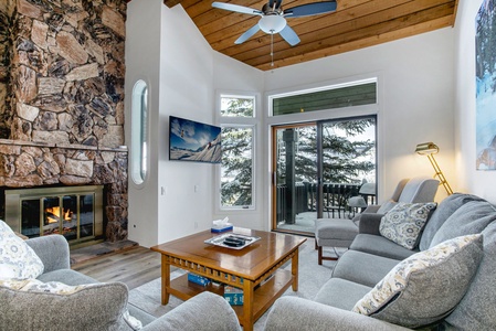 A cozy living room with a stone fireplace, grey sofas, a wooden coffee table, and a wall-mounted TV. Large windows and a sliding door open to an outdoor view, letting in natural light.