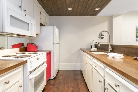 A small kitchen with white cabinets, a white stove, a refrigerator, a microwave, a sink with a flexible faucet, a red toaster, and wooden countertops and floor.