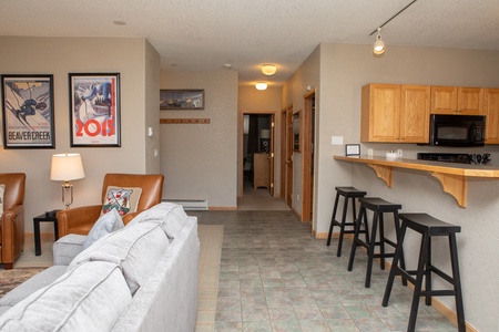 A modern, open-plan living area with light gray sofa, brown leather chair, bar with three black stools, kitchen with wooden cabinets on the right, and posters on a beige wall to the left.