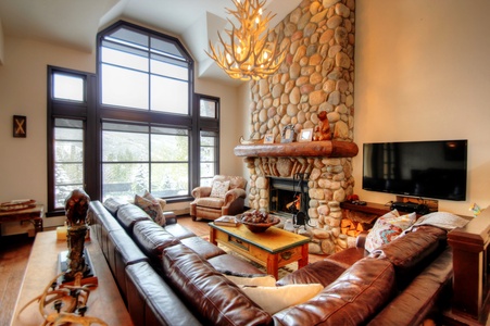 A cozy living room with a large stone fireplace, leather sectional sofa, armchair, wooden coffee table, and a mounted flat-screen TV. High ceilings and large windows let in natural light.