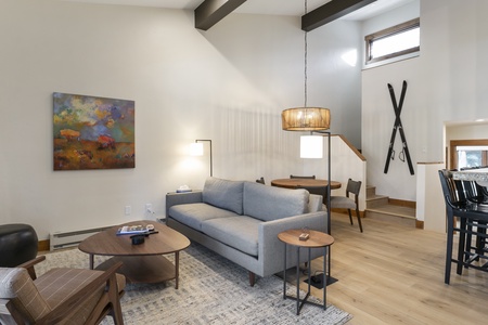 A modern living room features a gray sofa, wooden coffee table, wall art, and pendant light. A staircase leads to an upper level, and a pair of skis is mounted on the wall.