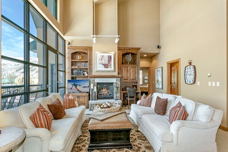 A spacious living room with high ceilings featuring two white sofas, a large ottoman, a fireplace, wooden bookshelves, and large windows with a view of snowy mountains.