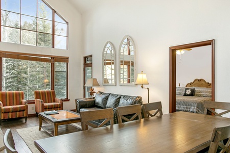 A living room with high ceilings features a dining table, couches, and armchairs near large windows. A doorway opens to a bedroom with a bed. Several lamps illuminate the space.