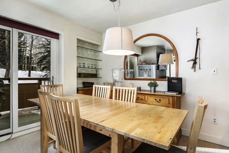 A dining area with a wooden table and six chairs. Features a large wall mirror, pendant light, and sliding glass door leading outside. Cabinets with glassware and a plant on a wooden sideboard.