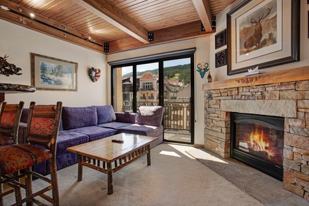 A cozy living room with a purple sectional sofa, rustic wooden coffee table, stone fireplace, and large glass doors opening to a balcony with mountain views. Various artworks adorn the walls.