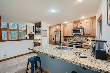 A modern kitchen with wooden cabinets, stainless steel appliances, granite countertops, an island with a sink and two stools, and a coffee maker on the countertop. Large windows are visible in the background.