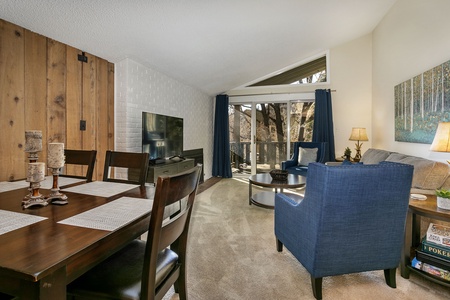 A cozy living and dining area with a wooden dining table, blue armchair, gray sofa, and a TV. Large window with blue curtains provides natural light. Decor includes lamps, candles, and artwork.