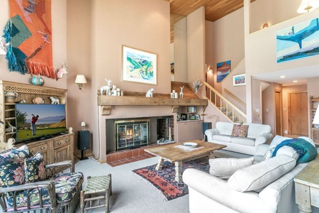 Living room with high ceiling featuring a fireplace, TV, and assorted furniture. Stairs lead to an upper level. Walls are adorned with various artworks and decorative items.