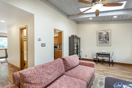 Living room with a ceiling fan, skylight, and a brown sofa. A dining table with chairs is in the corner, and a doorway leads to a kitchen area. A cabinet and framed picture are visible.