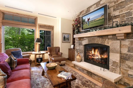 A cozy living room features a stone fireplace, a TV mounted above it showing a golf scene, a red sofa, an armchair, a wooden coffee table, and large windows with a green outdoor view.