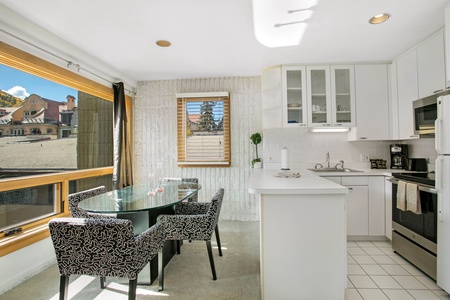 Bright kitchen with white cabinets and countertops, a small dining table with black patterned chairs, large window with a view of buildings, and a window with blinds.