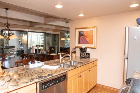 A modern kitchen with marble countertops, a double sink, a dishwasher, and a coffee maker. A dining area with a glass table is visible in the background. A framed picture hangs on the beige wall.