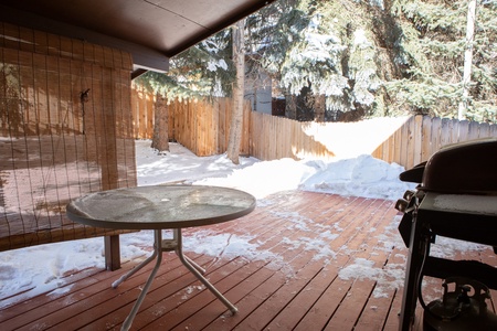 A snow-covered patio with a round glass table and a barbecue grill on a wooden deck. A wooden fence and tall evergreen trees are in the background.