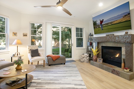 A cozy living room with a TV above a fireplace showing a golfer. The room has a round coffee table, a cushioned chair with a footrest, and large windows with a view of a garden.