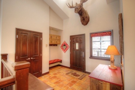 An entryway featuring a wooden front door, a bench with storage, a colorful rug, a side table with a lamp, and a mounted moose head above the door.