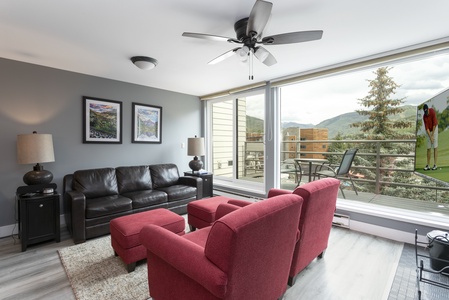 Living room with a leather sofa, two red chairs, a ceiling fan, and large windows overlooking a balcony with a mountain view. Two framed artworks are on the wall.