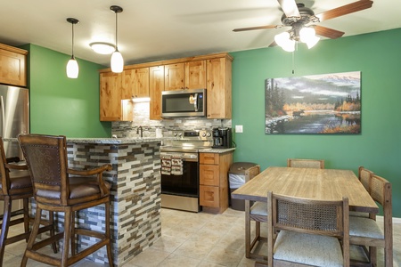 A kitchen with a breakfast bar, wood cabinets, stainless steel appliances, and a dining area. The bar has two chairs, and a landscape painting decorates the green wall. A ceiling fan is overhead.