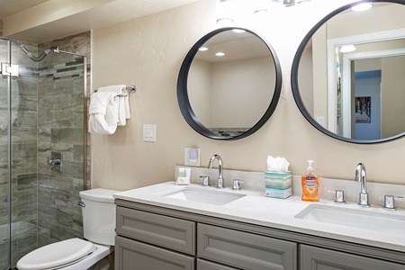 A bathroom with a double sink vanity, circular mirrors, and a glass-enclosed shower. The countertop holds a tissue box and hand soap dispenser. Towels are hanging beside the toilet.