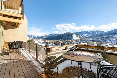 A snowy mountain view from a wooden deck with metal furniture, a fire pit table, and a barbecue grill. The clear sky and sunlight illuminate the scenic landscape.