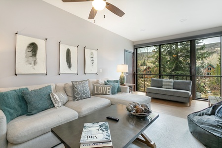 Modern living room with light gray sectional sofa, teal and gray cushions, framed feather artwork on the wall, large windows showcasing a wooded area outside, and a coffee table with magazines and decor.