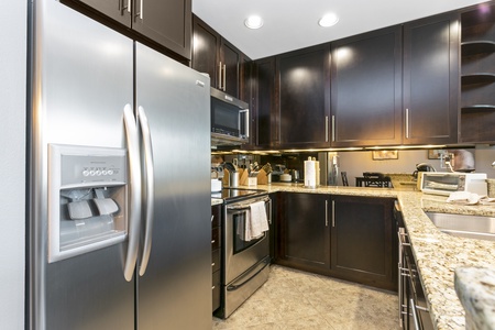 Modern kitchen with stainless steel refrigerator, dark cabinets, and granite countertops. Electric stove, sink, toaster, and knife set are visible.