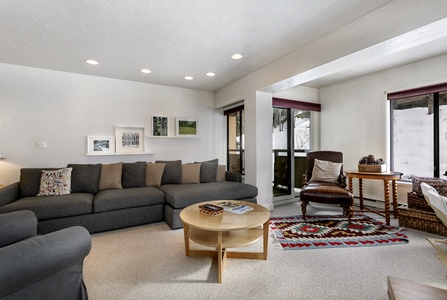 A modern living room features a gray sectional sofa, a round wooden coffee table, framed artwork on white walls, an armchair with a leather cushion, and a window with a snowy view outside.