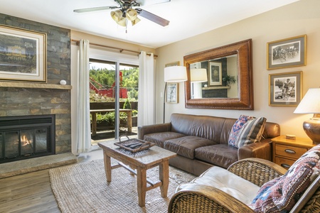 A cozy living room features a leather sofa, a wicker chair, a wooden coffee table, and a fireplace. A large mirror and framed photos adorn the walls. Sliding glass doors lead to an outdoor patio.