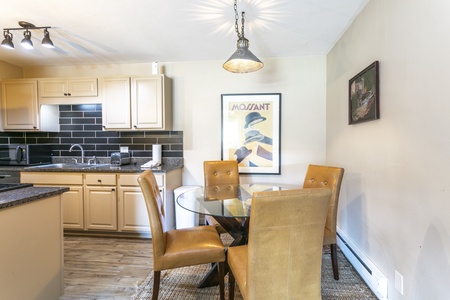 A modern kitchen with beige cabinets, black tile backsplash, and a round glass dining table with four brown leather chairs. A framed art poster and a hanging light fixture are visible on the walls.