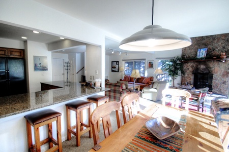 A spacious open-concept living area with a kitchen counter featuring wooden bar stools, a dining table with a pendant lamp, and a cozy living room with a stone fireplace and large windows.