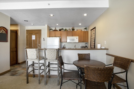 A kitchen and dining area featuring a bar with four high chairs, a round table with four wicker chairs, wooden cabinets, a microwave, a stove, decorations on top of the cabinets, and a wooden door.