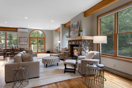 A cozy living room with a stone fireplace, beige sofas, a circular ottoman, and large windows. A dining area is visible in the background.