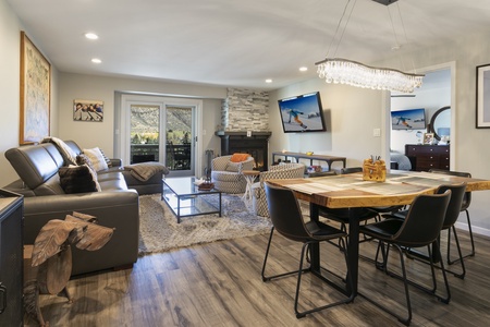 Modern living and dining area with wood floors, a gray sofa, TV, fireplace, dining table with black chairs, and a sliding door to a balcony with mountain views.