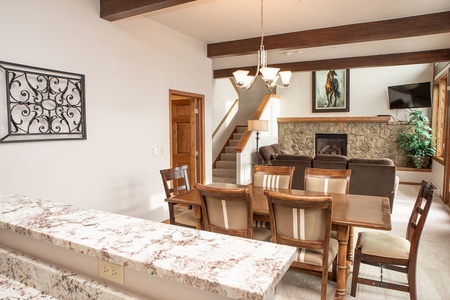 A living and dining area with wooden furniture, a stone fireplace, and a decorative wall hanging. A staircase leads upstairs, and a painting and TV are mounted on the walls.