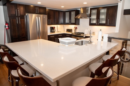 A modern kitchen with a large white island, brown cabinets, stainless steel appliances, a sink, and six brown padded chairs around the island. The counters hold small kitchen appliances and accessories.