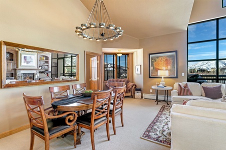 A dining area with a wooden table and chairs is set in an open living space. Large windows show a mountain view. The room features a chandelier, mirror, armchairs, and a couch with decorative pillows.