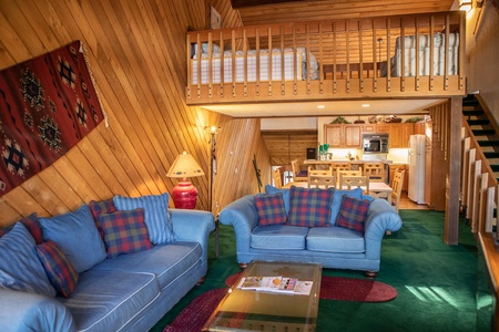 A cozy living area with blue couches, patterned pillows, a glass coffee table, and a red lamp. Wooden walls, a loft with a bed, and a kitchen with wooden cabinets and white appliances are visible.