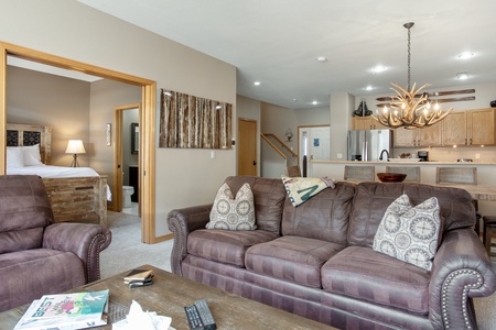 A living room with brown leather sofas, a coffee table, and a view of a kitchen and dining area with antler chandelier. An open doorway reveals a bedroom with a wooden bed and a bathroom.