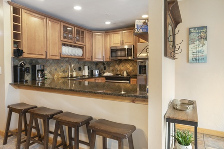 A kitchen with wooden cabinets, a granite countertop, stainless steel appliances, and a high counter with four wooden stools. A sign on the wall reads, “Welcome to our Chalet.”.