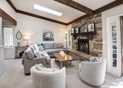 A cozy living room with a gray sectional sofa, two round chairs, a wooden coffee table, and a stone fireplace. The room features exposed wooden beams and a skylight, creating a warm, inviting atmosphere.