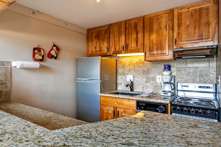 A small kitchen with wooden cabinets, a refrigerator, stove, coffee maker, and granite countertops. Two mitts hang on the wall beside a paper towel holder.