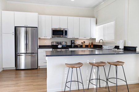 Modern kitchen with white cabinets, stainless steel appliances, a dark countertop, and a wooden floor. Three wooden stools are positioned at a peninsula under a window with blinds.