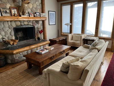 A cozy living room features a stone fireplace, a beige couch, a wooden coffee table, an armchair, and framed pictures on the mantle. Large windows provide natural light, and the floor is wooden.